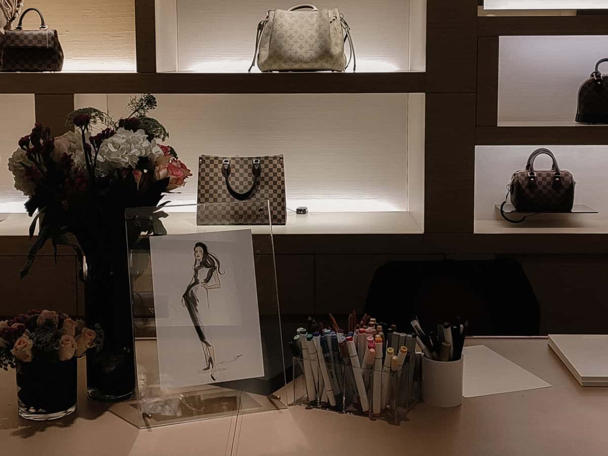 Photo of an artist's set up for live event sketching at an in store Louis Vuitton event. The setup has an acrylic frame with as sketch, holders with art markers and flower arrangements with LV bags in the background. Artist BY HANLI